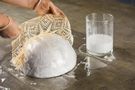 Next to a glass containing a glue-water mixture, gloved hands place the soaked lace doily over the polystyrene mould.