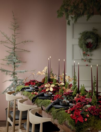 Table festive avec un décor forestier composé de poinsettias, de mousse, de pommes de pin et de bougies devant une porte avec une couronne.