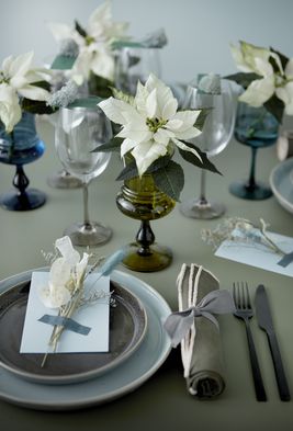 Mesa puesta con platos, cubiertos, vasos, servilletas y poinsettias blancas en recipientes de cristal.