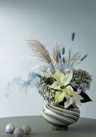 Arrangement of cream poinsettias and blue dried flowers in vase with Christmas baubles next to it
