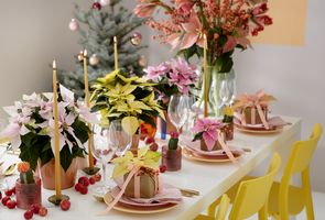 Table avec chaises jaunes et centre de table coloré composé de poinsettias, pommes, colis, bougies et cactus.